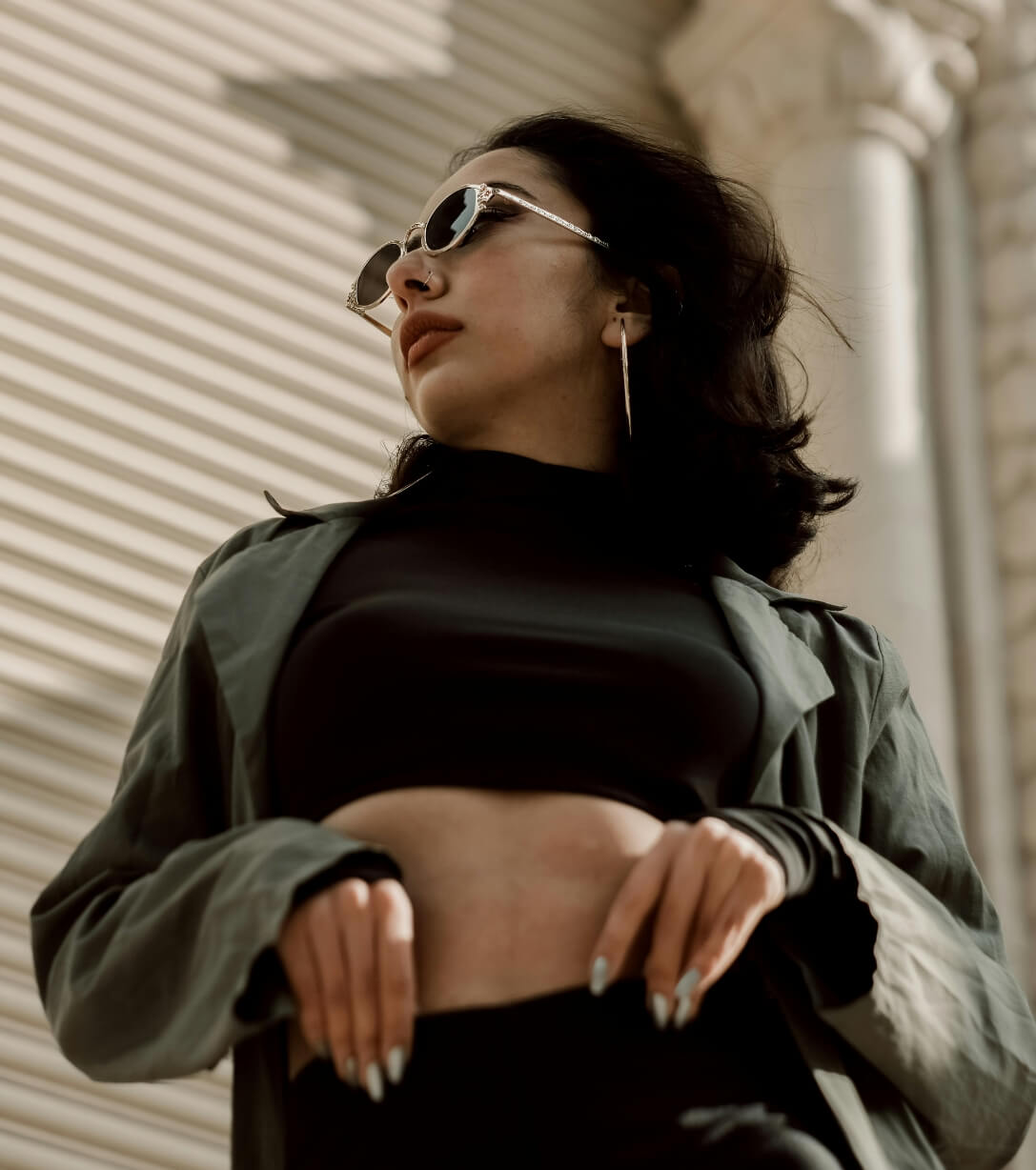 young adult woman wearing cropped turtle neck top, army green jacket, black sunglasses posing with her hands on her front waist
